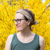 A women stands in front of a bright yellow flowering bush wearing glasses and a rolled up bandana in her hair.